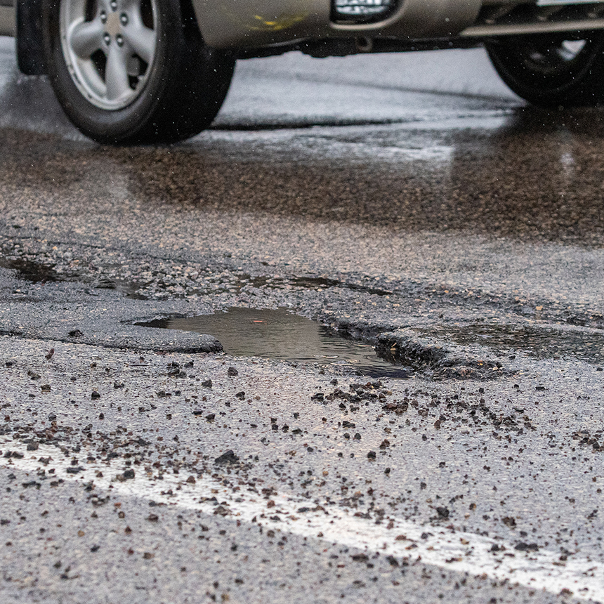 Close-up of a pothole on a wet road with a car tire in the background